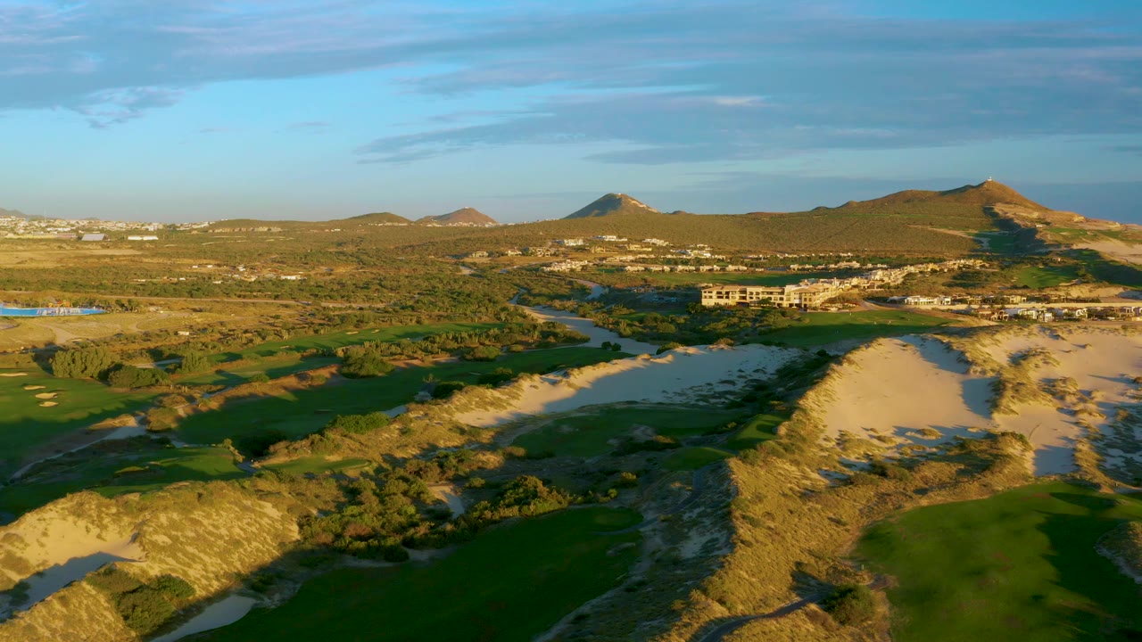 Wide aerial frame over Diamante Cabo San Lucas with sandy dunes in the foreground, green fields around the river, distant hillside buildings, blue sky, and no visible people.