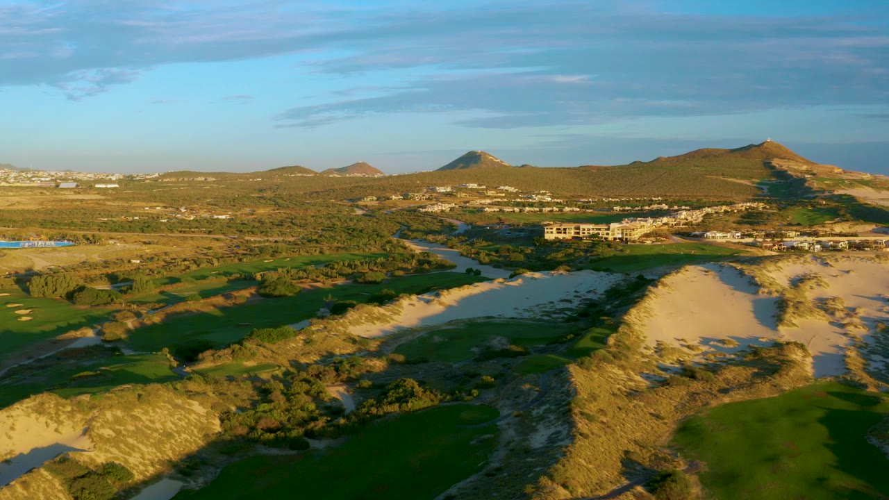 Mid-clip aerial frame in Diamante Cabo San Lucas showing dunes and scrub in front, green fields and river vegetation beyond, distant structures, mountains, and no visible people.