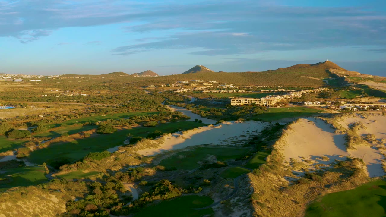 Wide aerial frame over Diamante Cabo San Lucas with river water curving through green terrain, sandy banks, low buildings, distant hills, warm sunlight, and no visible people.