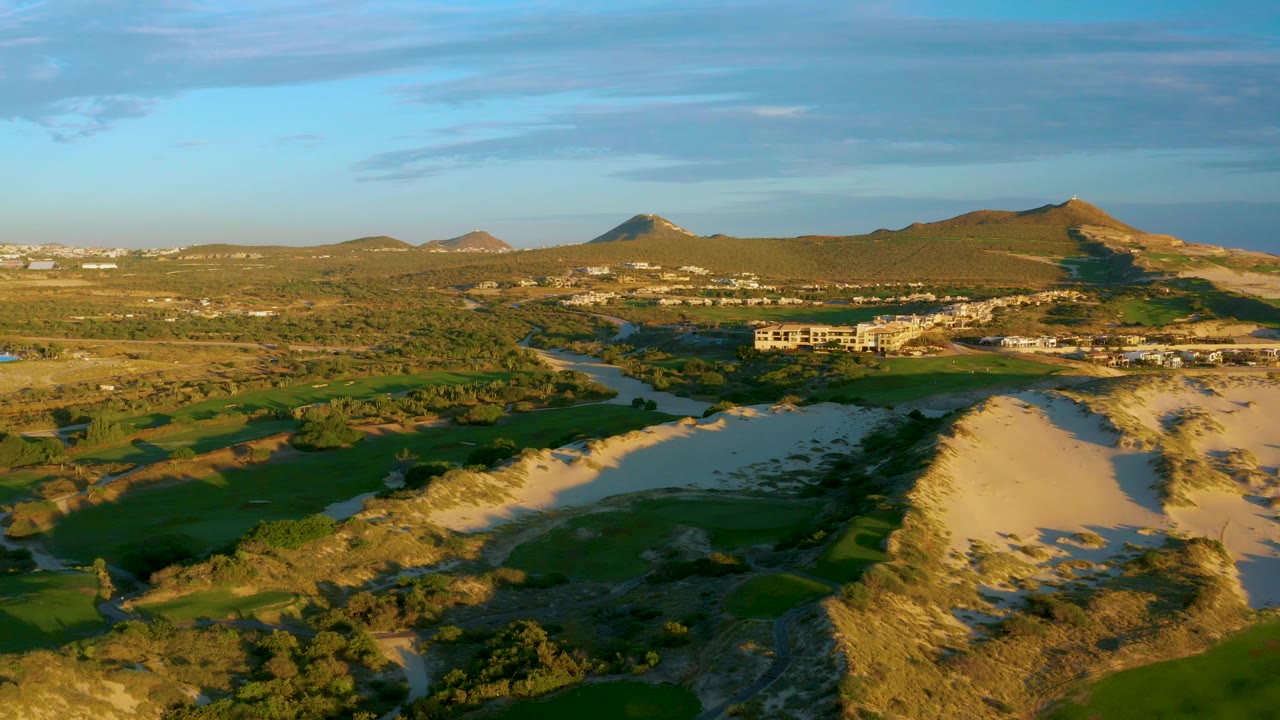 Diamante Cabo San Lucas aerial landscape with a river running past green fairway-like fields and sandy dunes, with a built complex near the center and no visible people.