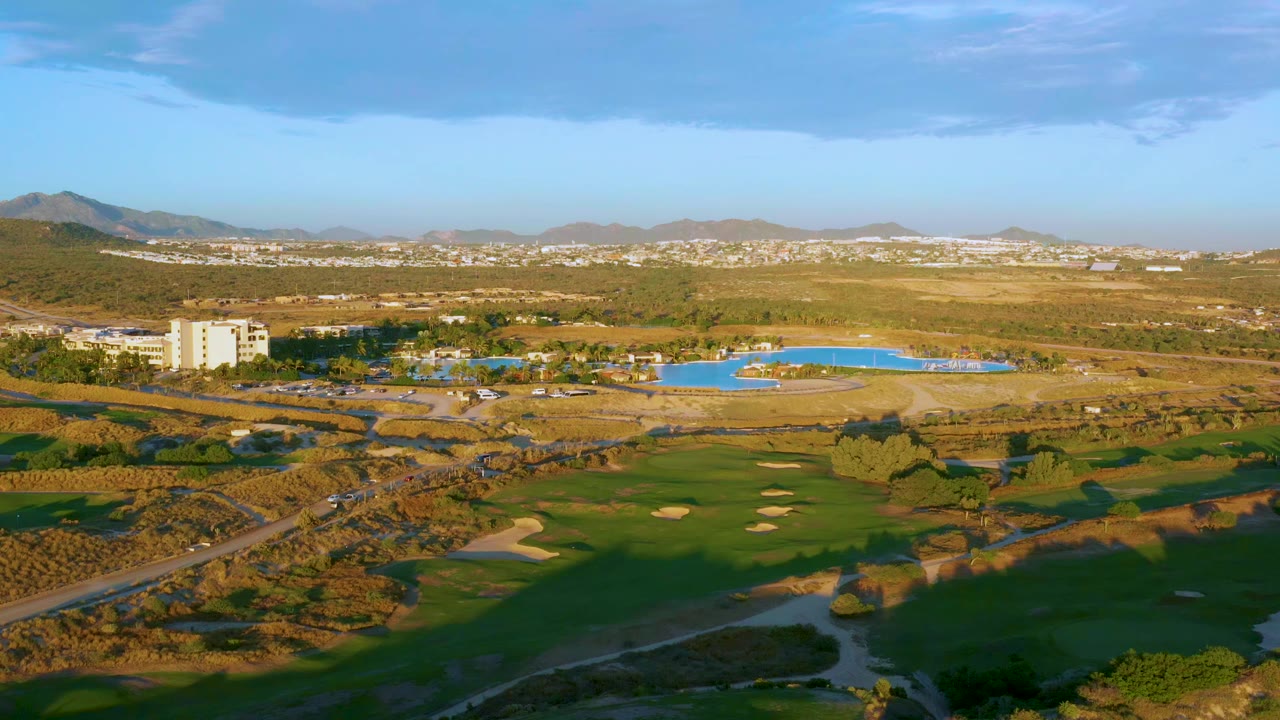 Second aerial frame at Diamante Cabo San Lucas with the road and golf greens lower in frame, the bright resort pool centered beyond them, distant hills on the horizon, and no visible people.