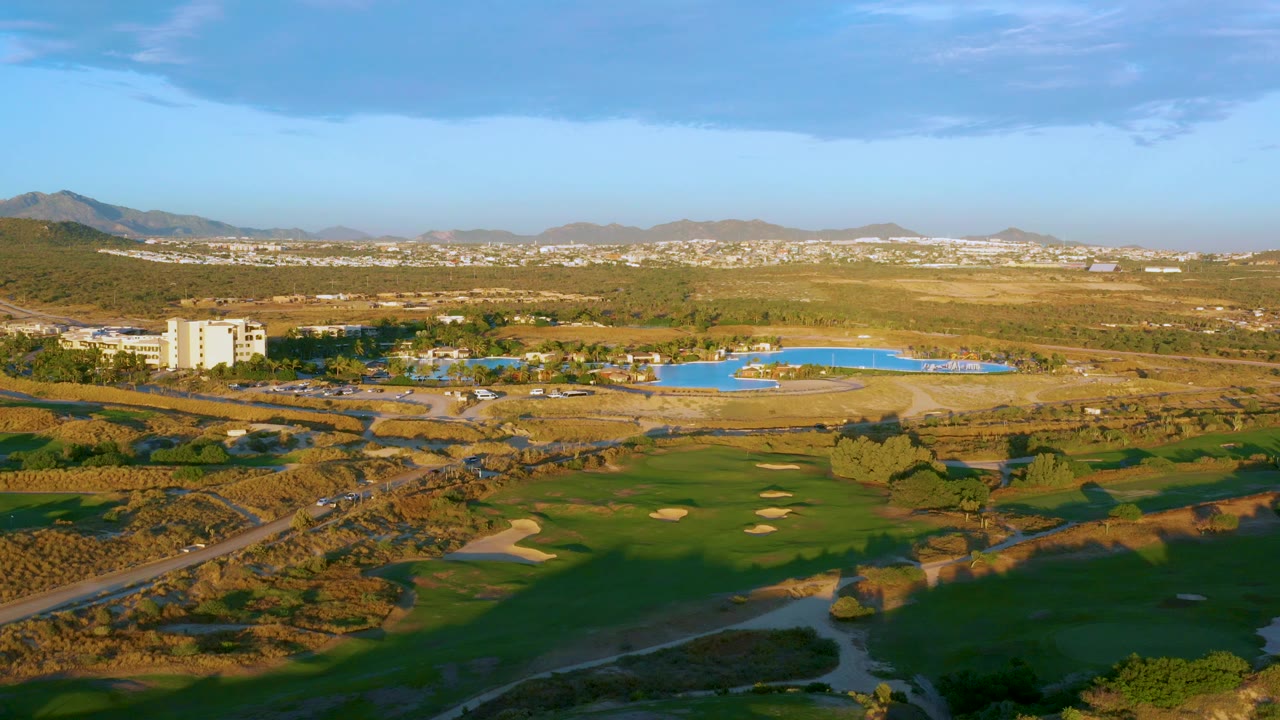 Mid-clip aerial view at Diamante Cabo San Lucas showing the resort building on the left, blue pool across the midground, golf fairways and bunkers below, brown terrain beyond, and no visible people.
