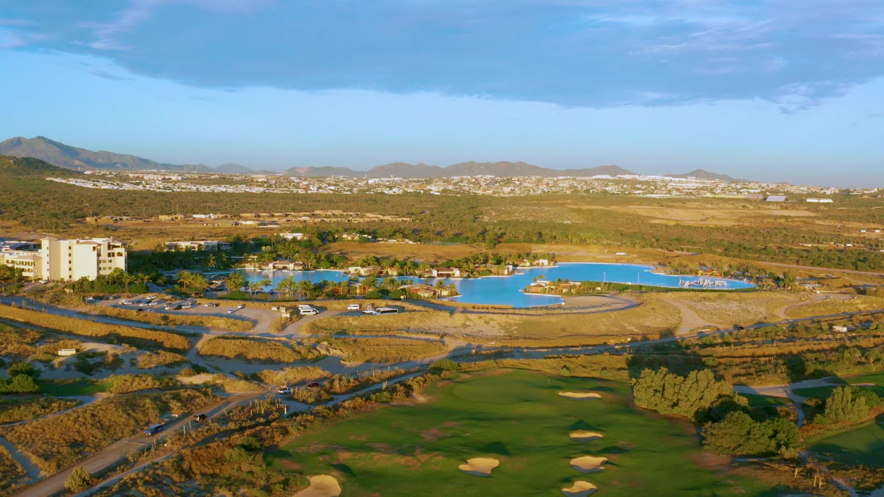 Elevated aerial view over Diamante Cabo San Lucas with green fairways in the foreground, bright blue water across the midground, dry hills in the distance, and no visible people.