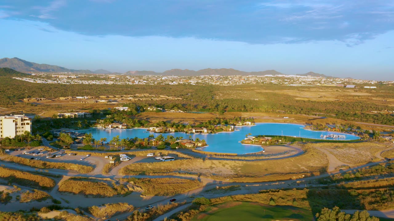 Opening aerial frame over Diamante Cabo San Lucas shows a bright blue lake bordered by dry grass, dirt roads, parked cars, distant buildings, mountains, and no visible people.