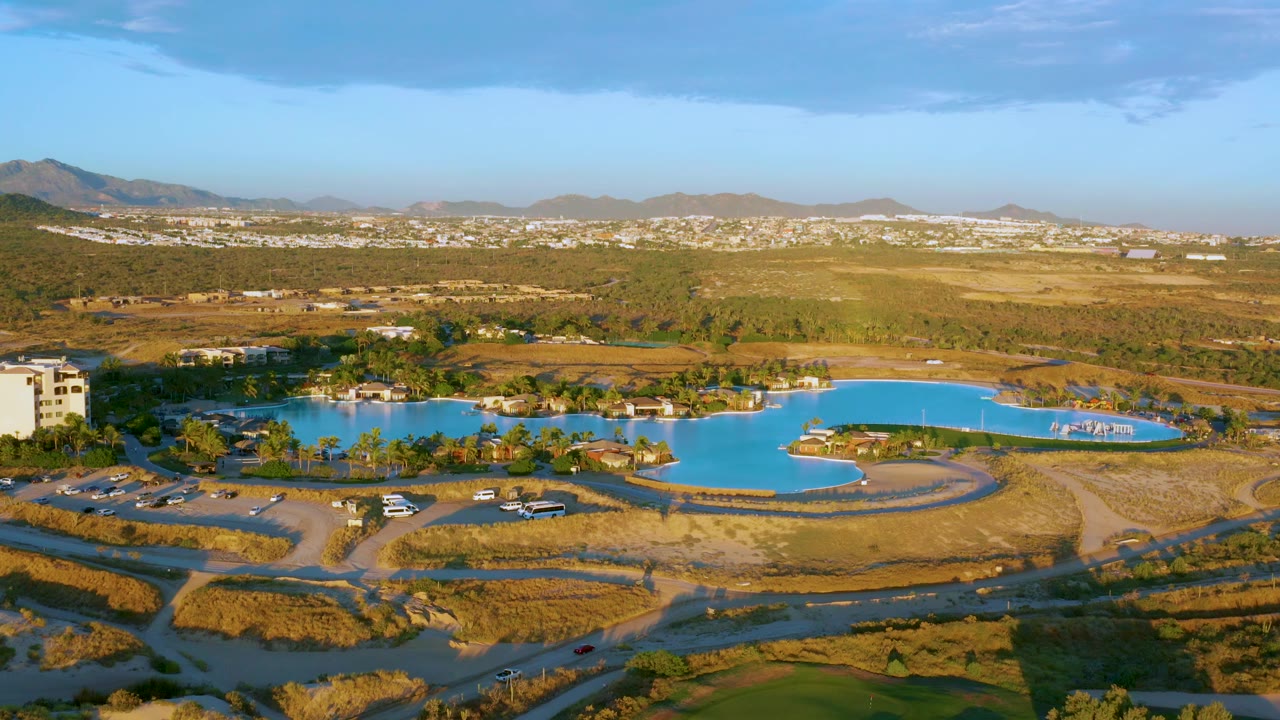 At 0:02, a nearby building at the lake edge is more apparent while the blue water, dry ground, roads, distant town, hills, and no visible people remain in view.