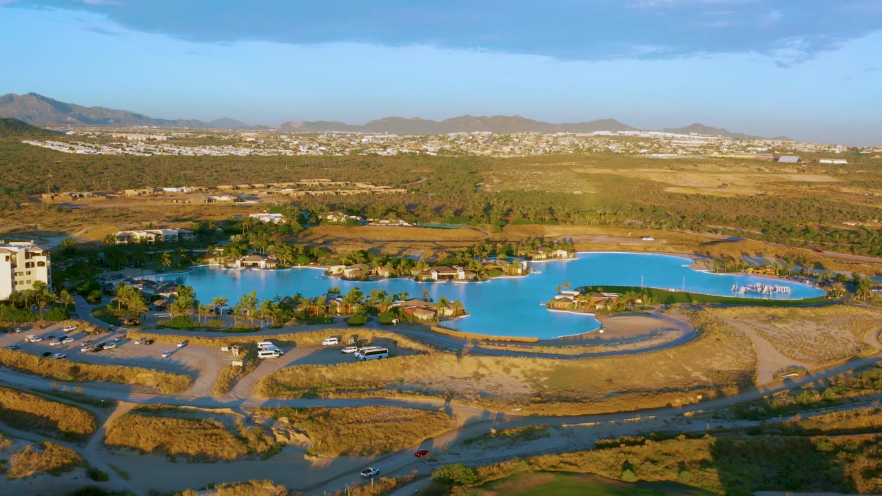 Aerial lake scene with dry grass foreground, parked vehicles, buildings, town, and mountains.