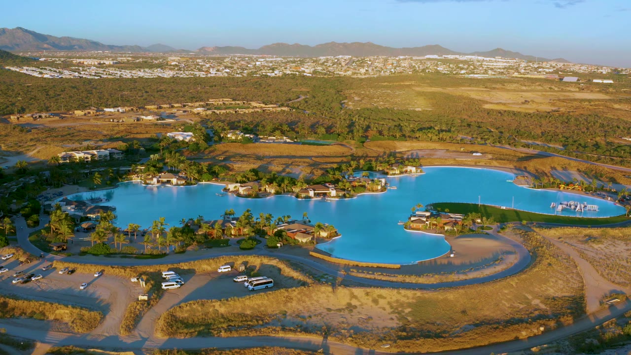 Wide drone view of lake water beside exposed ground and parking areas.