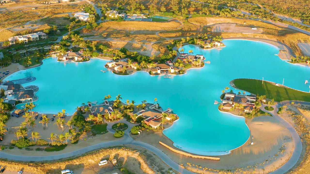 At 0:01, the drone view over Diamante Cabo San Lucas keeps the bright blue lake central, with homes, palms, curved roads, small vehicles, and no visible people.