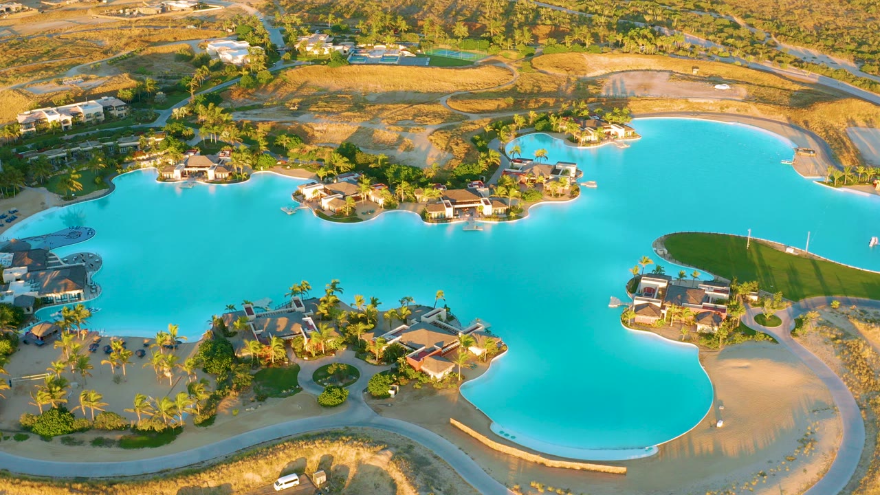 Aerial frame over Diamante Cabo San Lucas showing a turquoise lagoon, curved shorelines, villas, palm landscaping, dry surrounding land, and no visible people.