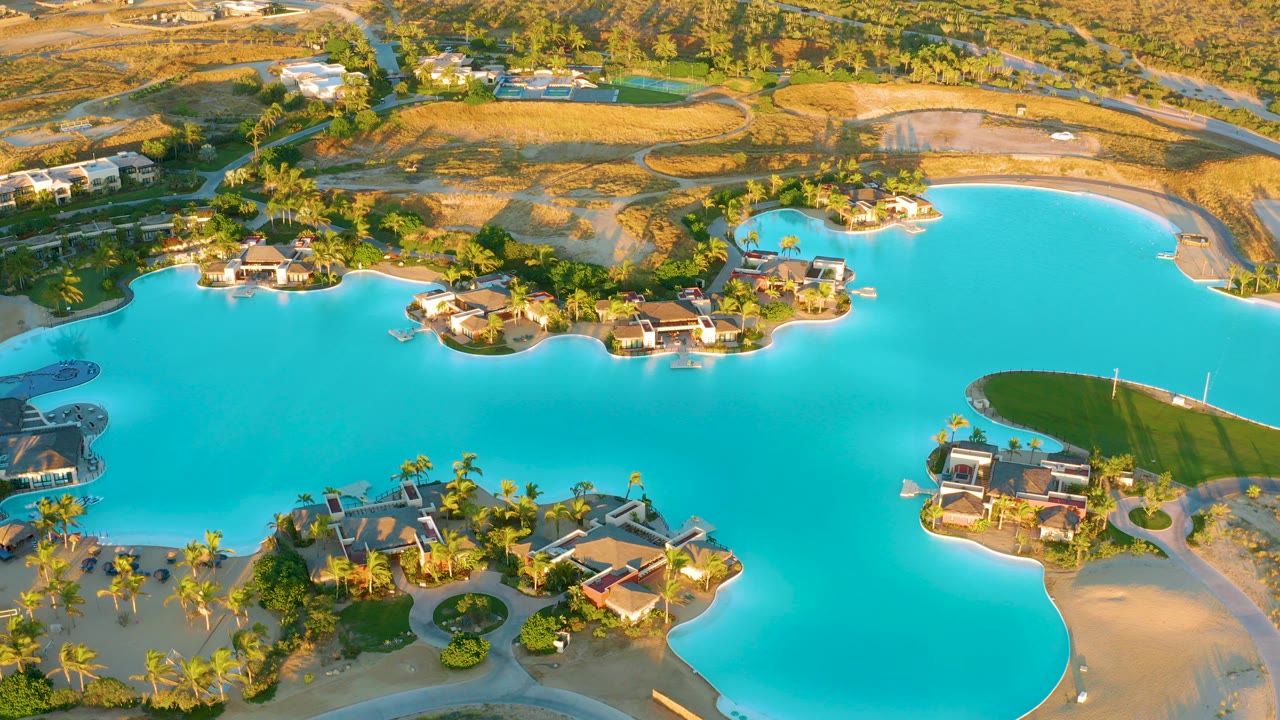 High aerial frame over Diamante Cabo San Lucas showing turquoise lake water, villa-style homes, palm trees, roads, dry surrounding land, and no visible people.