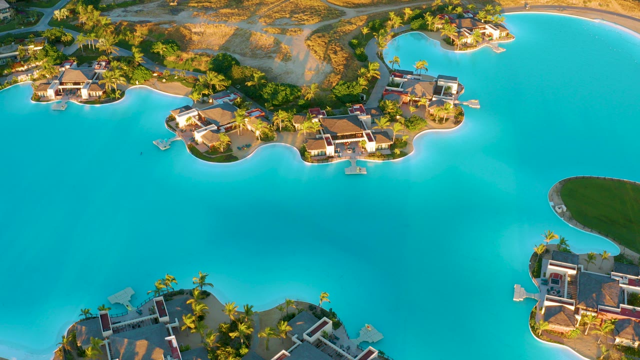High aerial frame over a bright turquoise lagoon at Diamante Cabo San Lucas, with waterfront villas on small curved islands, palms, sandy ground, and no visible people.