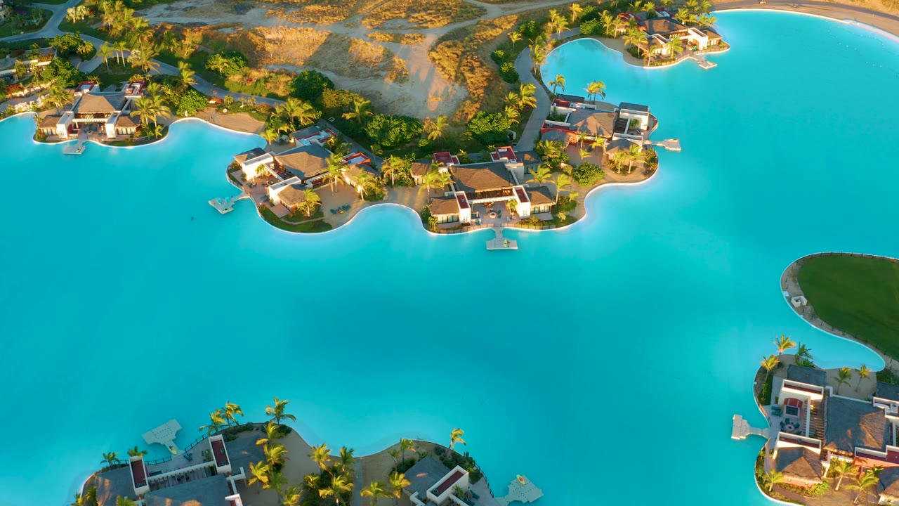 High drone frame over Diamante Cabo San Lucas showing a broad turquoise lagoon, waterfront houses on landscaped islands, a partial green landform at right, and no visible people.