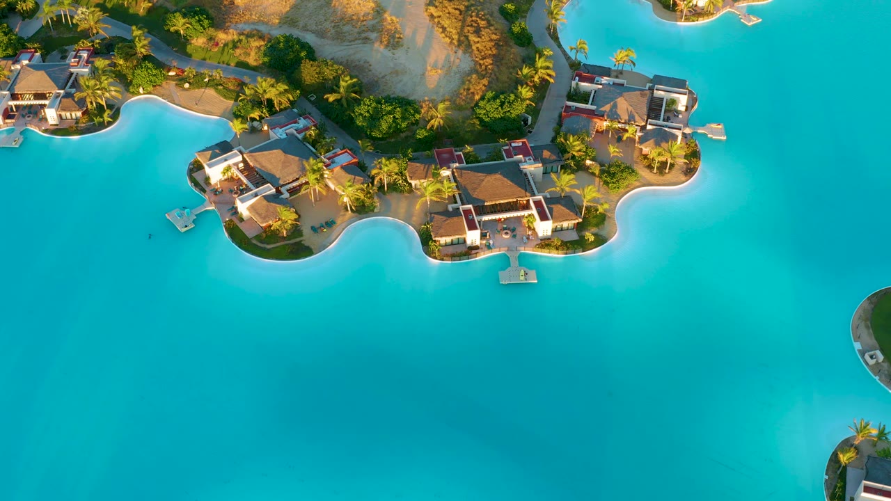 Drone view over Diamante Cabo San Lucas with villas and palm trees tracing a scalloped sandy shoreline above open turquoise water, with no visible people.