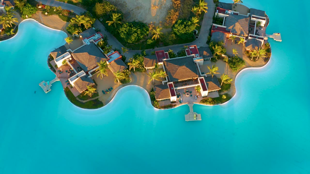 Top-down aerial frame of thatched overwater bungalows, palm trees, curved sand edges, small docks, and turquoise water in Diamante Cabo San Lucas, Mexico, with no visible people.