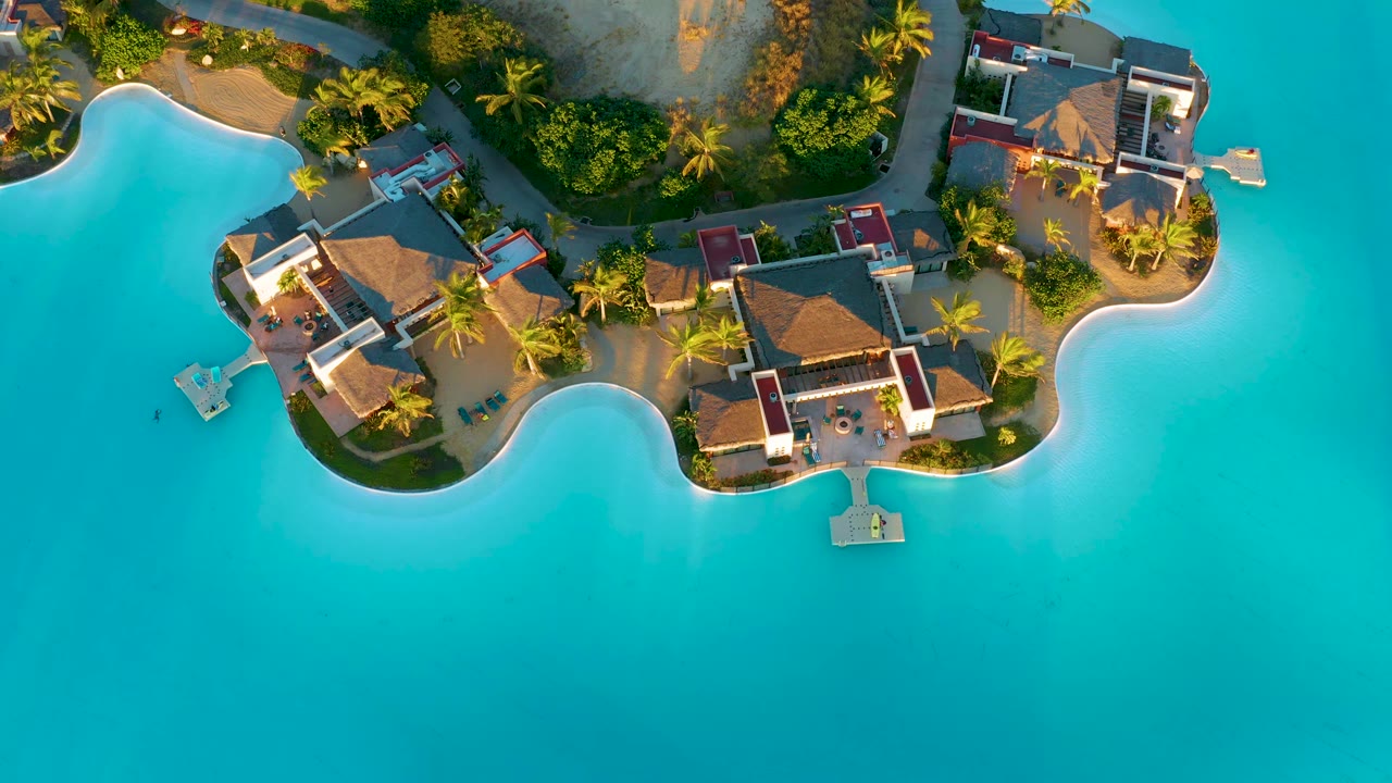 Aerial frame looking down on the central bungalow roofs and garden paths, with turquoise water spreading across the lower half and no visible people.