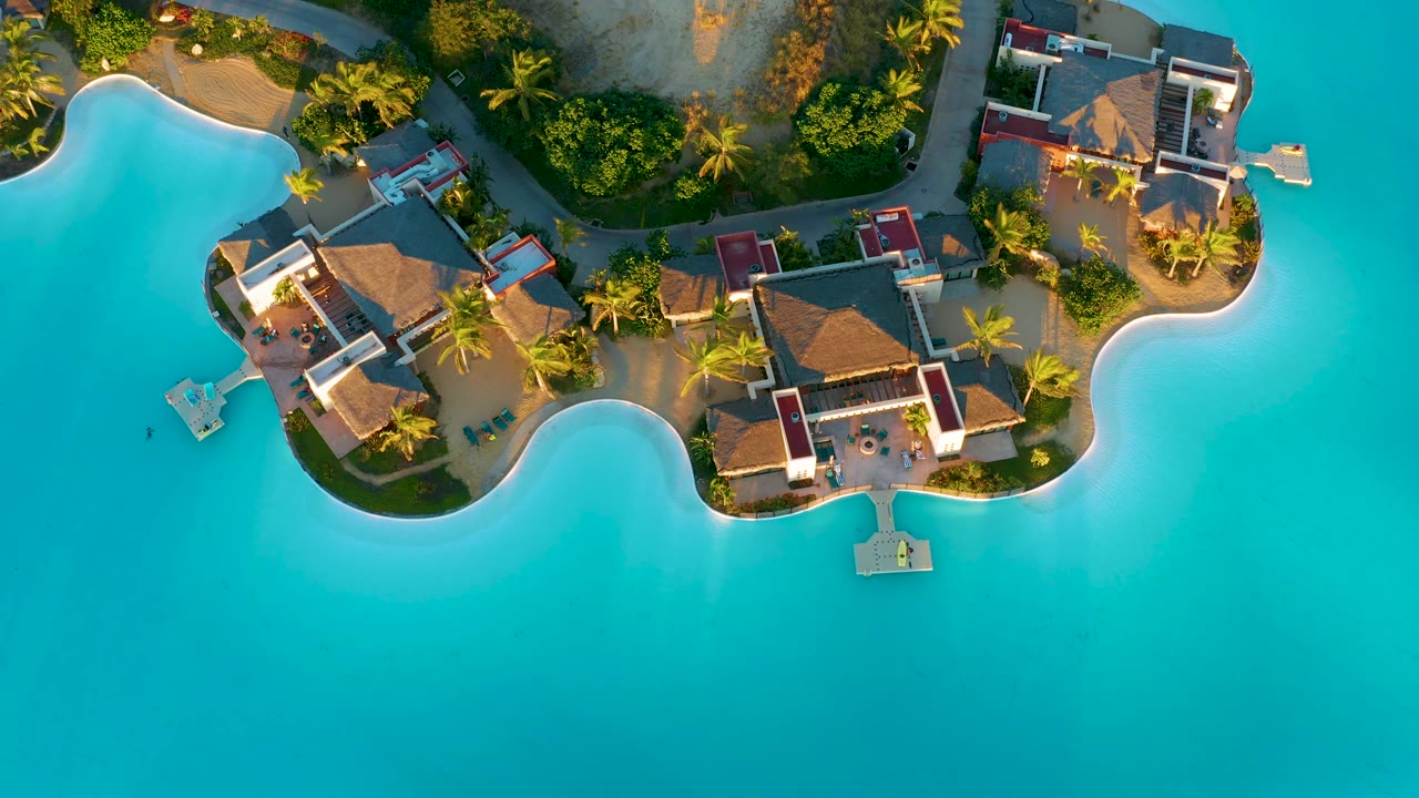 Overhead view of thatched bungalows, a curved pale shoreline, palms, small platforms, and clear turquoise water in Diamante Cabo San Lucas, Mexico, with no visible people.
