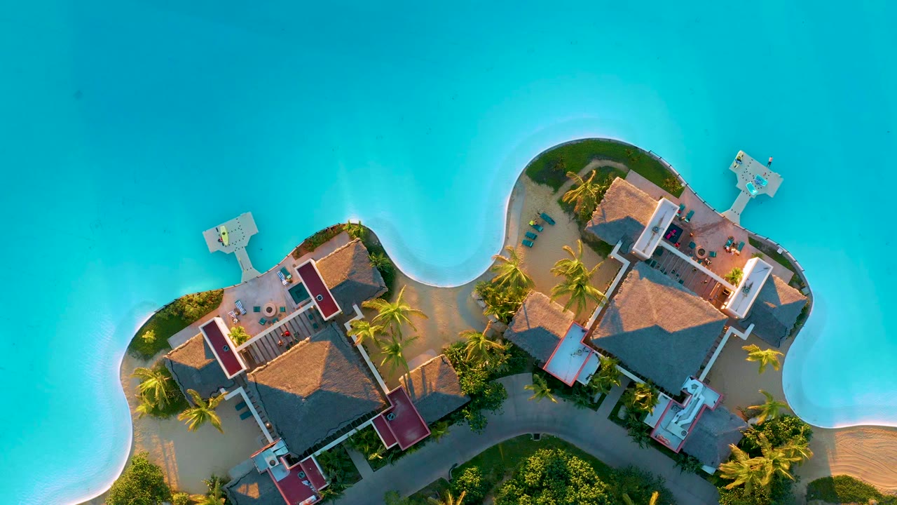 Overhead view at Diamante Cabo San Lucas showing resort buildings, palms, curved shoreline, small piers, turquoise water, and no visible people at the start of the clip.