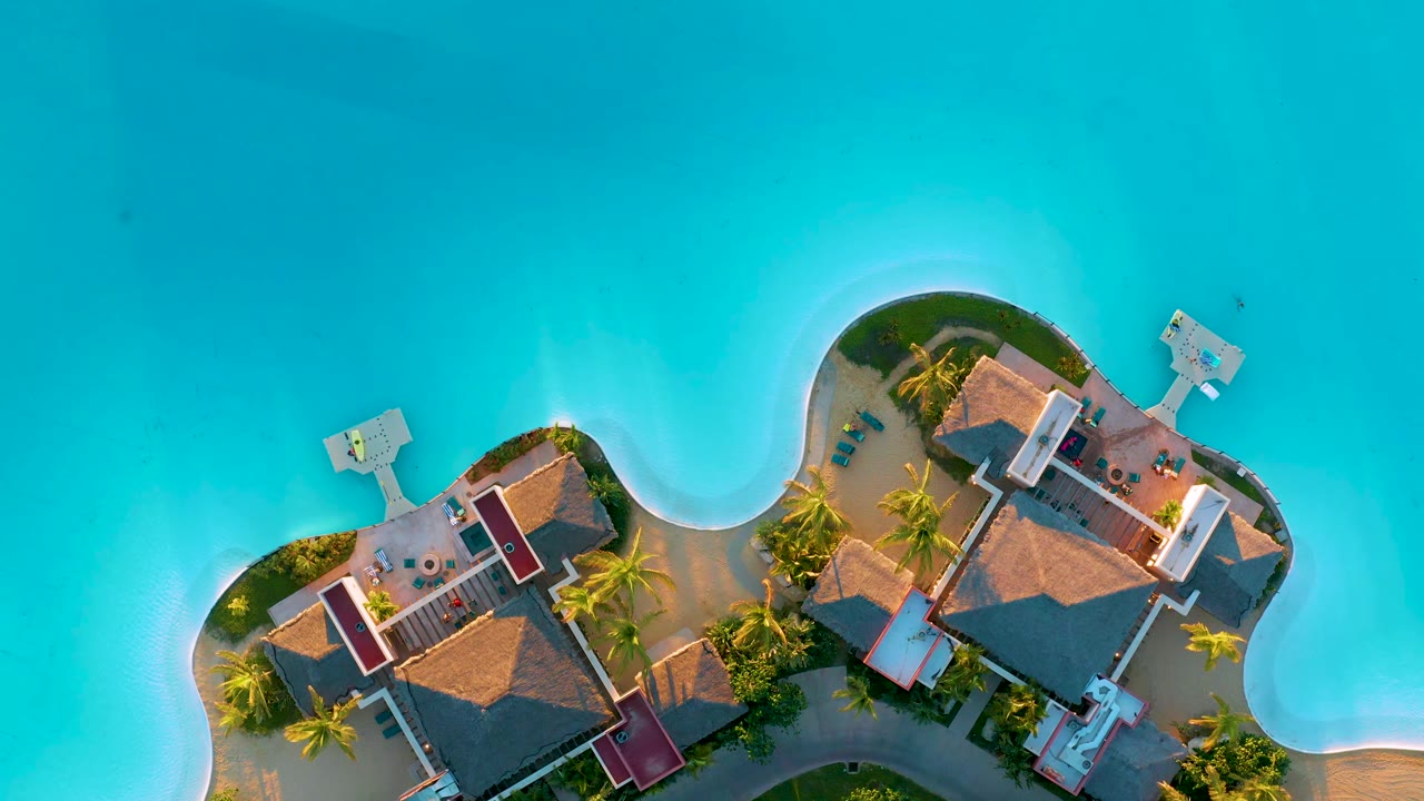 Straight-down aerial view in Diamante Cabo San Lucas with the island buildings slightly more centered below turquoise water, with docks and no visible people.