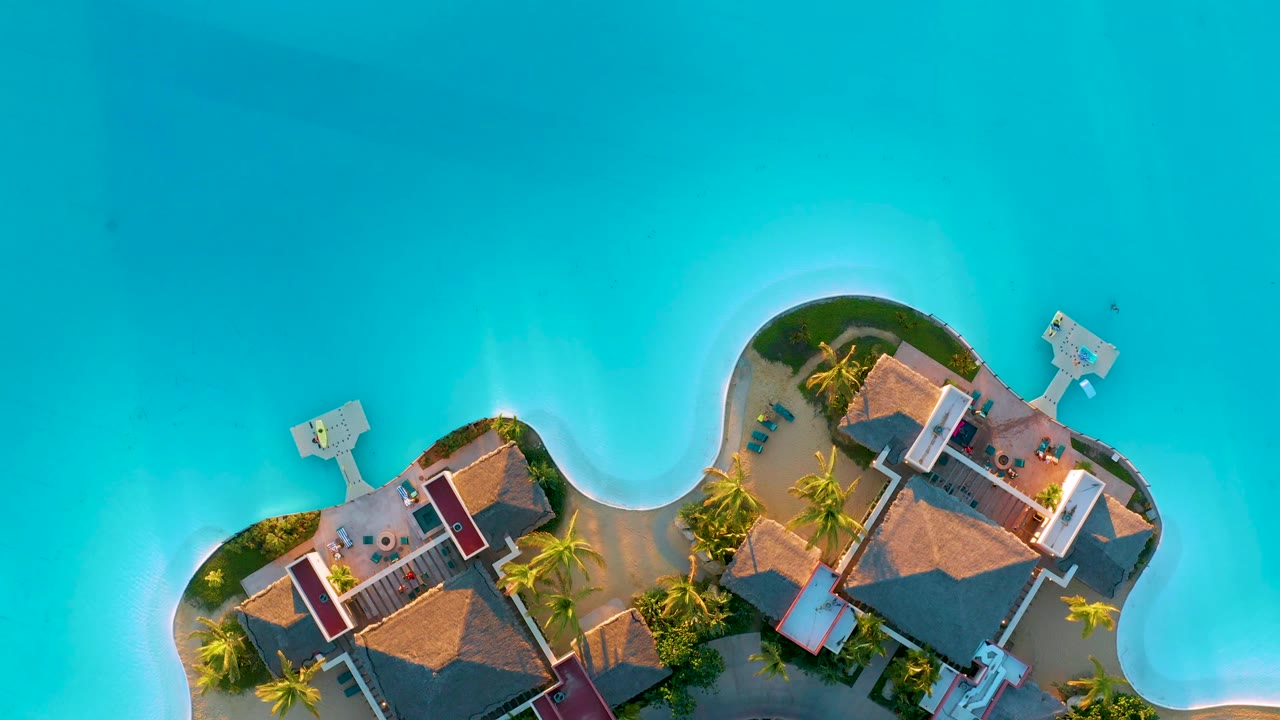 Overhead frame from Diamante Cabo San Lucas showing the curved island edge, thatched roofs, palm trees, small docks, clear turquoise water, and no visible people.