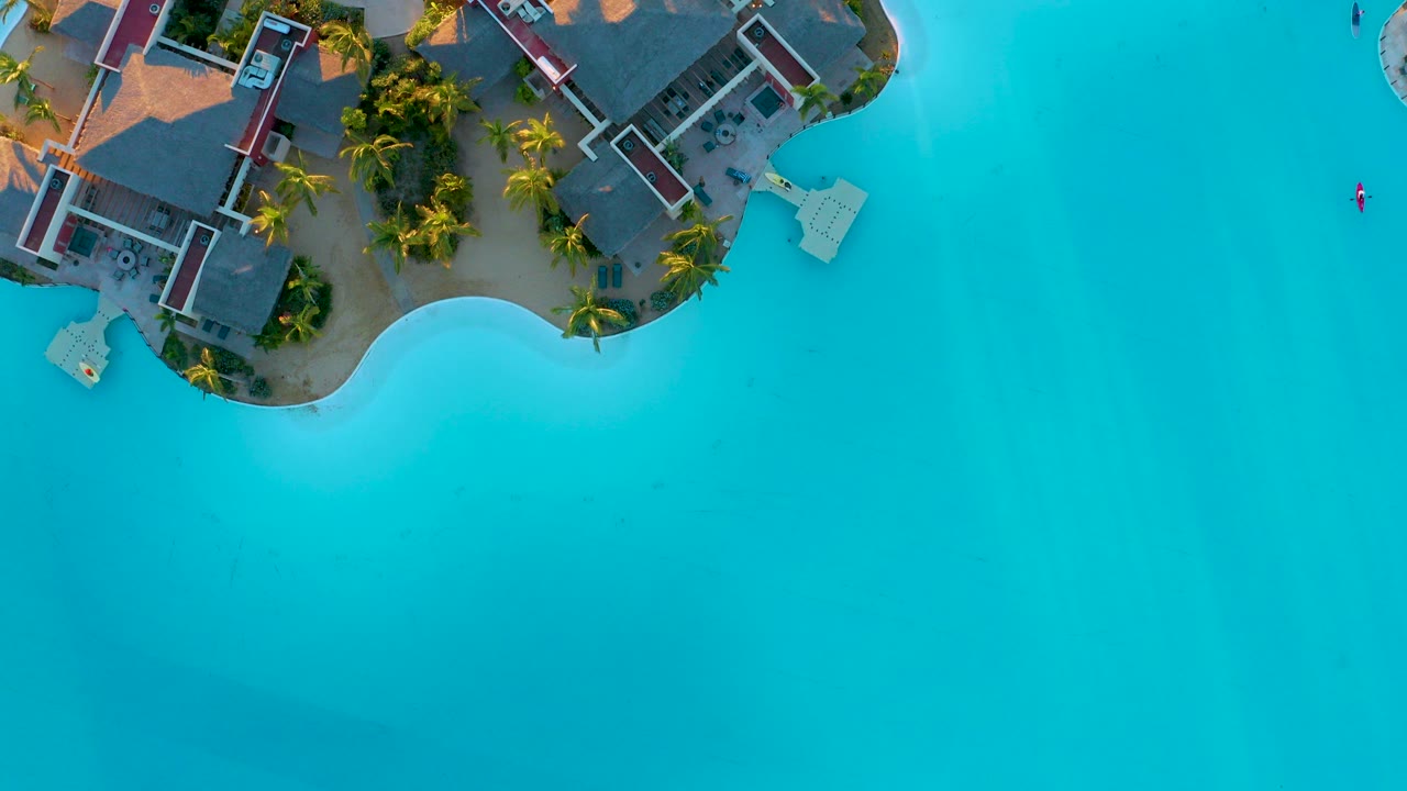 Overhead view in Diamante Cabo San Lucas showing resort rooftops, palm trees, sandy shoreline, and open turquoise water with no visible people.