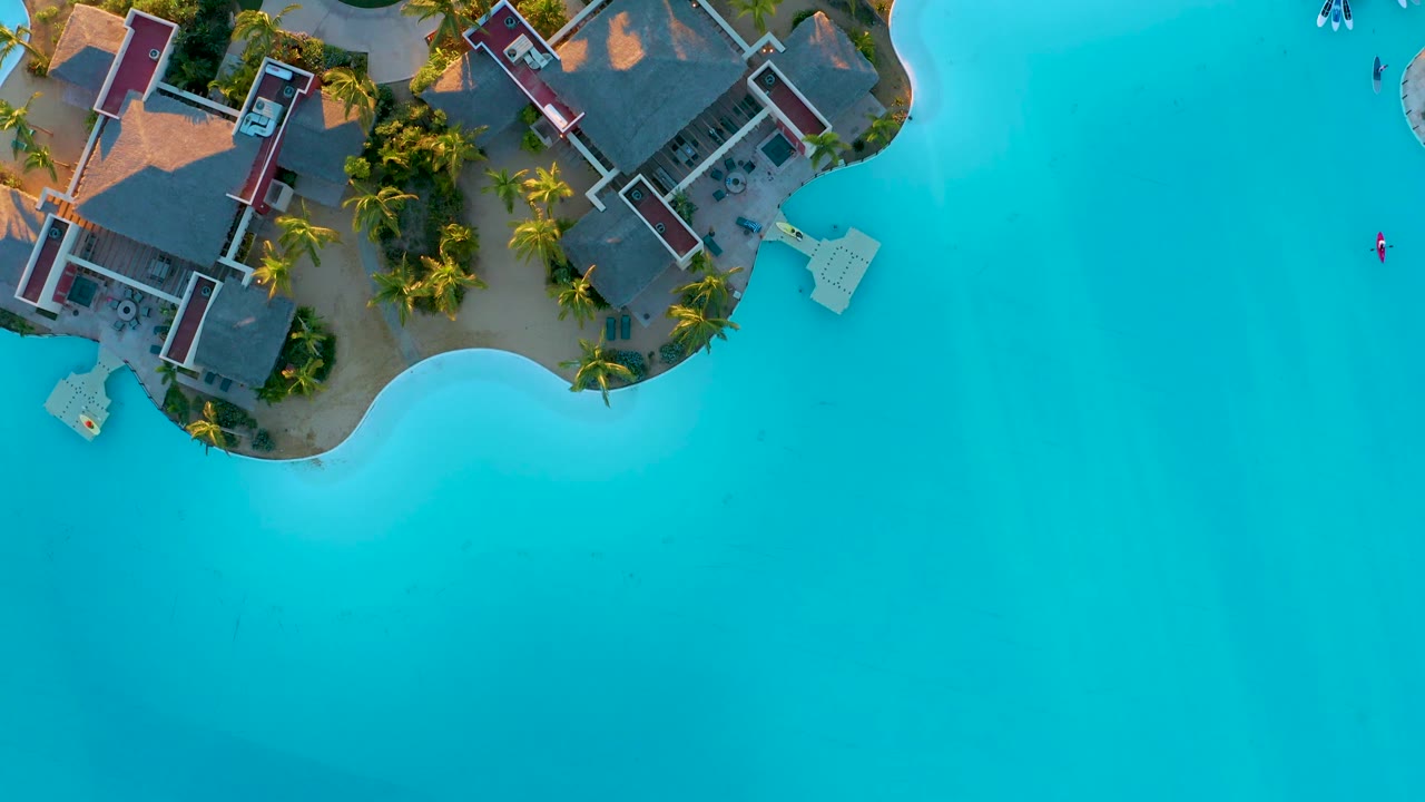 Top-down Diamante Cabo San Lucas frame showing resort buildings along the land edge, bright blue water, small boats near shore, and no visible people.