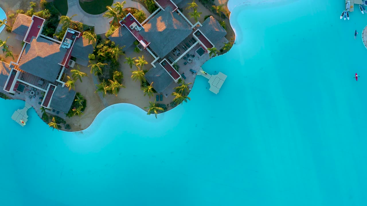 Drone view of resort rooftops, palms, and clear blue water.