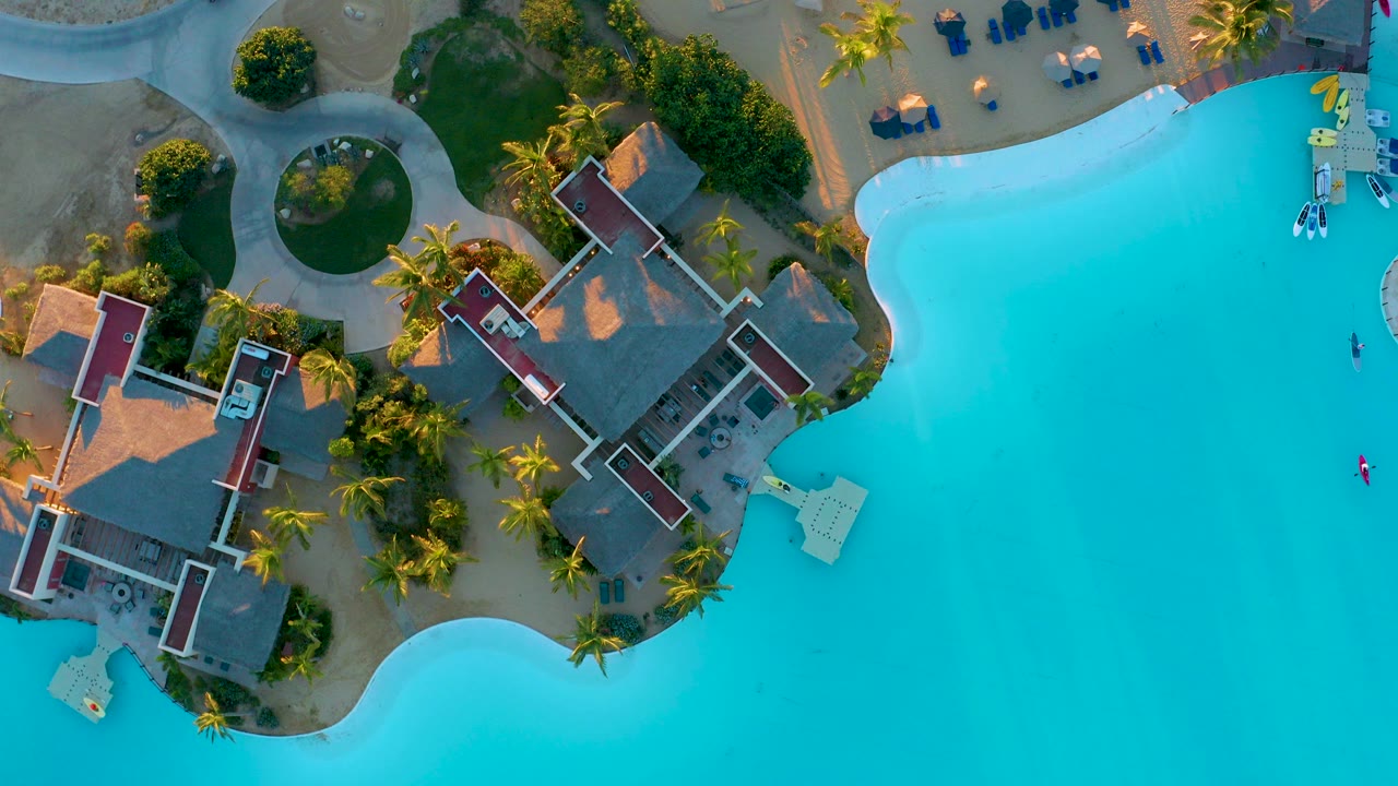 Overhead view of a bright blue resort pool, red-roofed buildings, palm trees, curved paths, and sandy grounds at Diamante Cabo San Lucas, with no visible people.