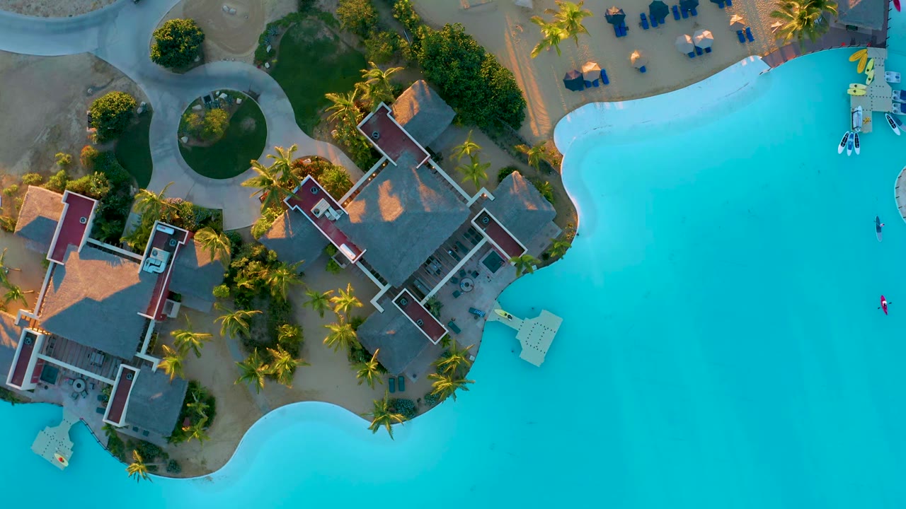 Aerial frame over the pool edge and central resort buildings at Diamante Cabo San Lucas, showing palms, paths, sand, and no visible people.