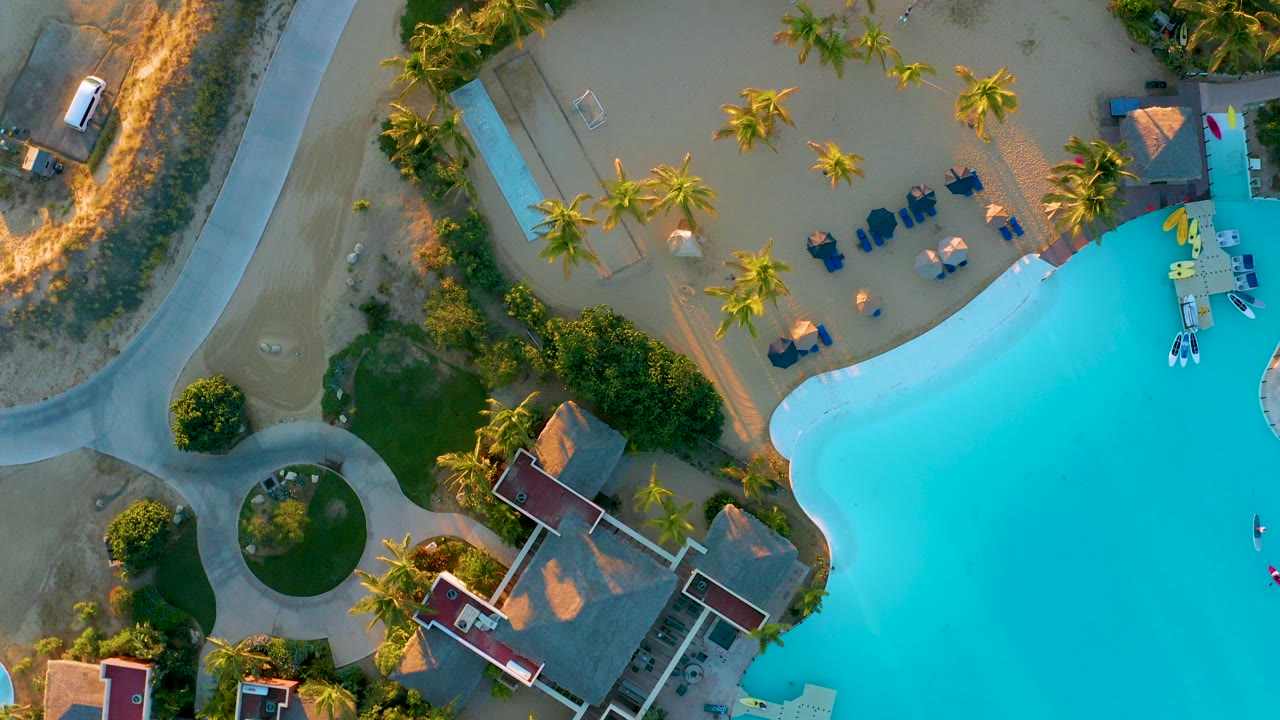 Top-down view of Diamante Cabo San Lucas resort grounds with turquoise pool water on the right, red-roof buildings below, curved paths, sandy landscaping, palm trees, and no visible people.