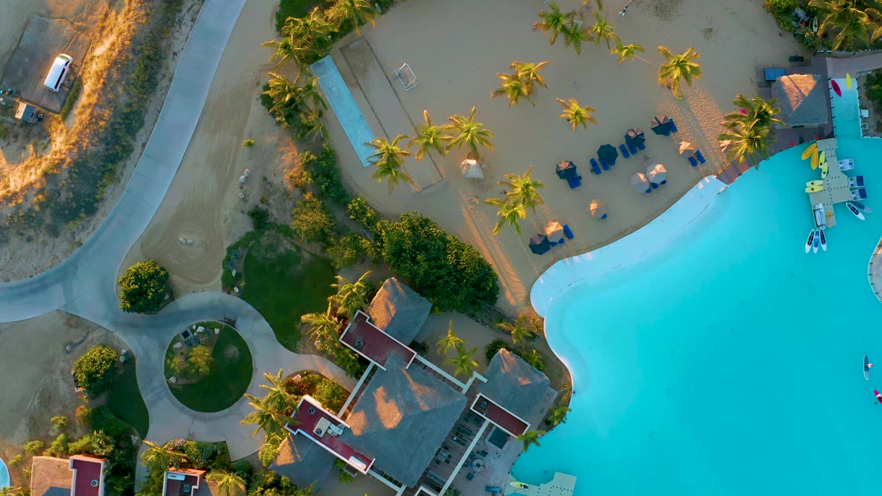 Overhead frame in Diamante Cabo San Lucas with bright pool water at right, palm trees across sandy grounds, lounge chairs near the pool, curved walkways, red-roof structures, and no visible people.
