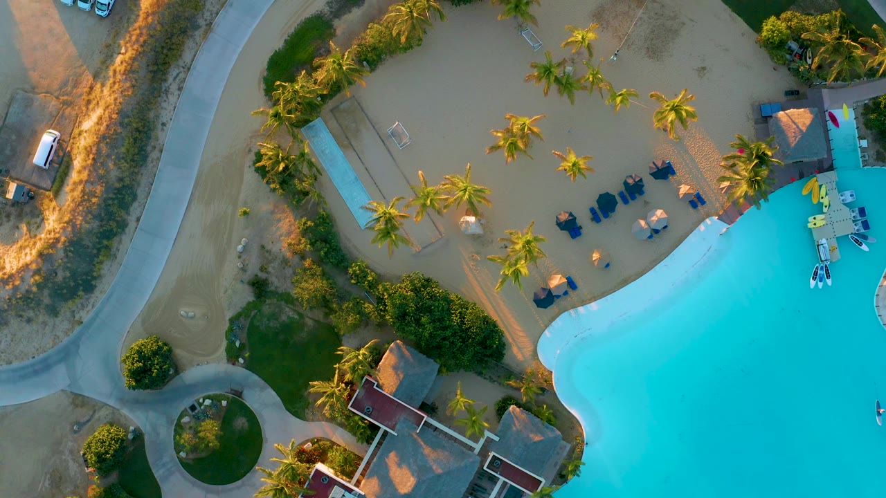 Overhead resort grounds with pool, palms, curved road, lounge chairs, and white cars.