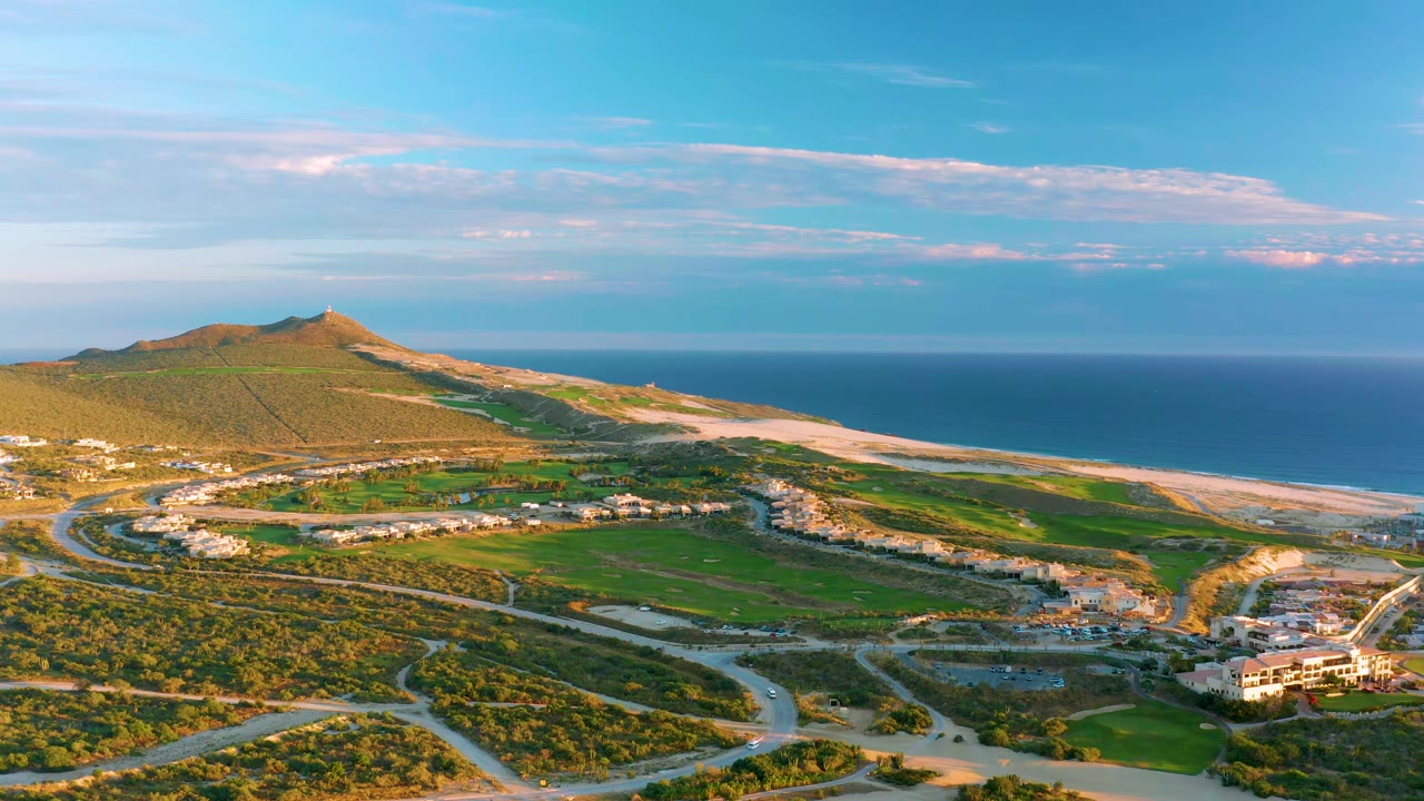 Wide drone view of hillside, beach, ocean, roads, fields, and lower-right building.