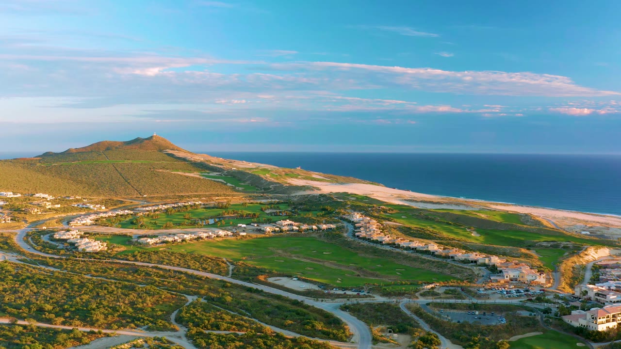 Aerial frame over Diamante Cabo San Lucas showing more of the road bends in the foreground, clustered low buildings, the sandy shoreline, blue water, and no visible people.