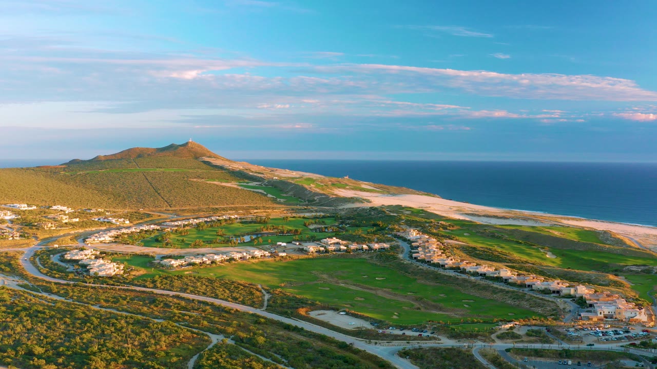 Opening aerial frame over Diamante Cabo San Lucas, Mexico, with a green hill at left, sandy beach and ocean at right, curved roads, homes, and no visible people.