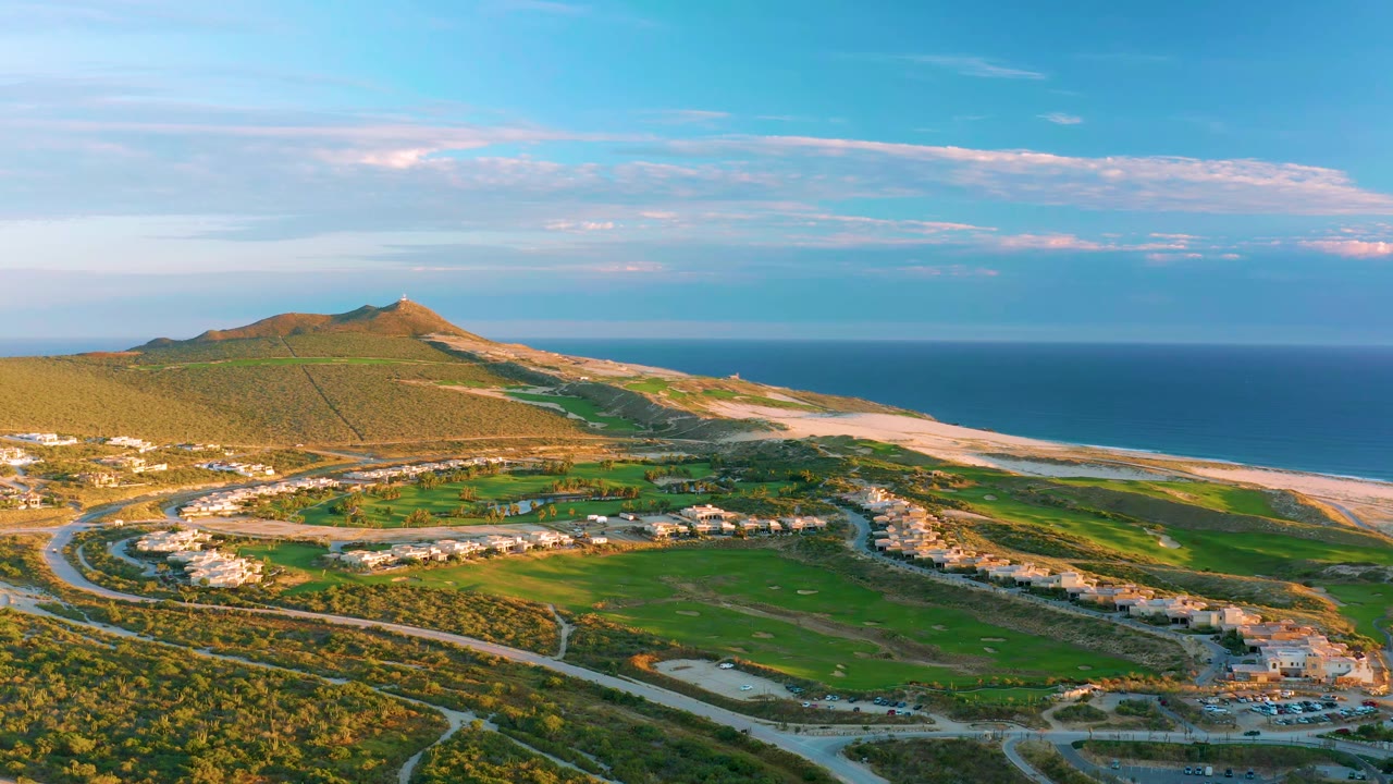 Wide aerial view in Diamante Cabo San Lucas with green fields below, a low coastal hill to the left, ocean beyond the sandy shoreline, and no visible people.