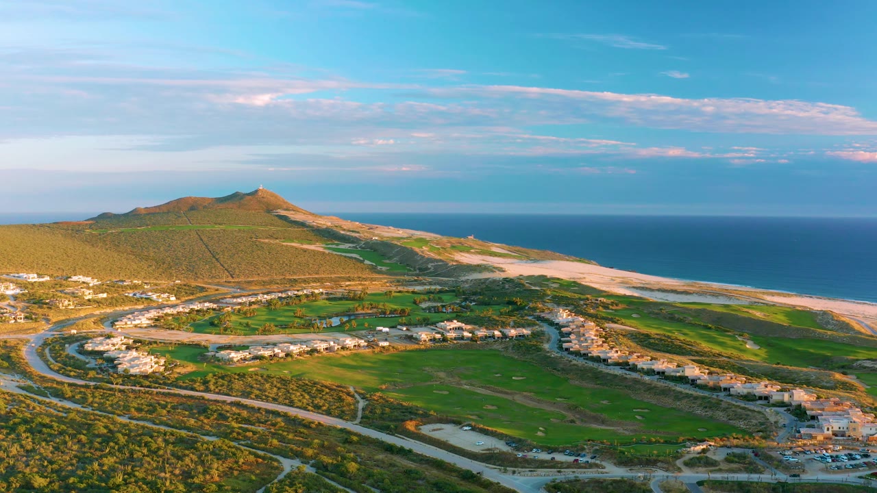 Aerial view over coastal roads, homes, green grass, hill, beach, and sea.