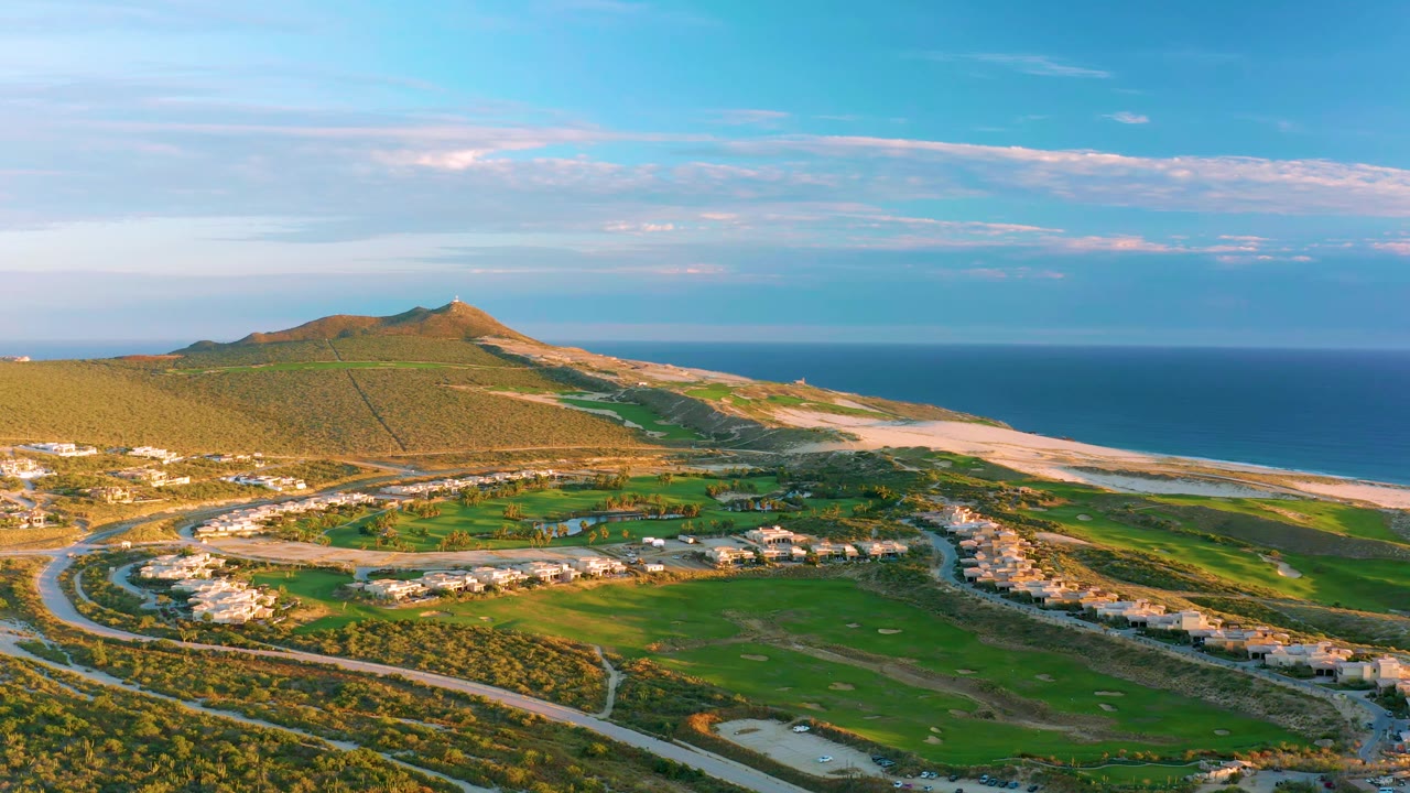Diamante Cabo San Lucas aerial frame with road loops and clustered residences below a low hillside, backed by a sandy beach, ocean horizon, and no visible people.