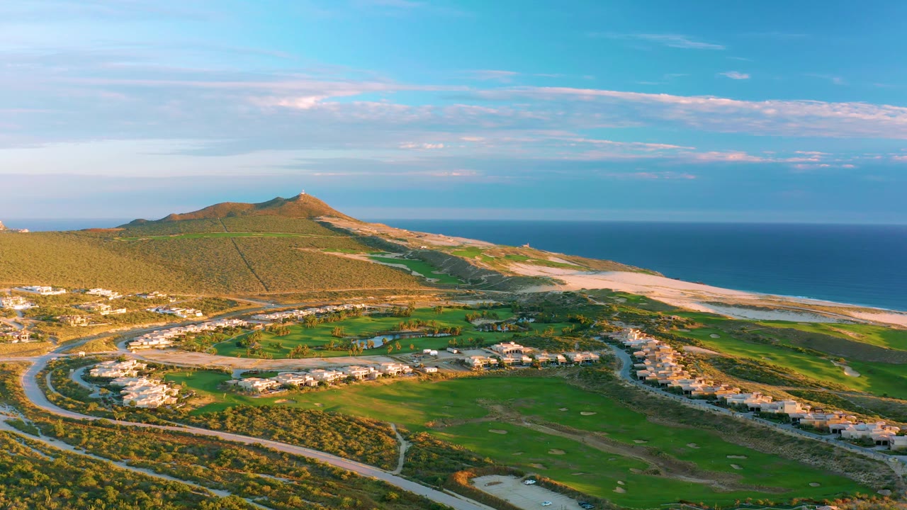 Drone view across coastal homes, green fairways, hill, beach, and ocean.