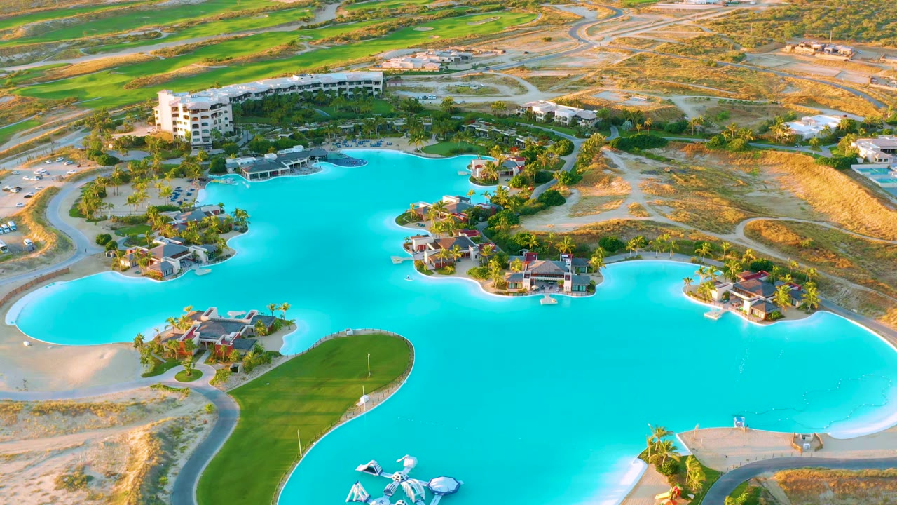Opening aerial frame over Diamante Cabo San Lucas showing turquoise lagoon water, island homes, a green peninsula, sandy roads, a multi-story building, and no visible people.
