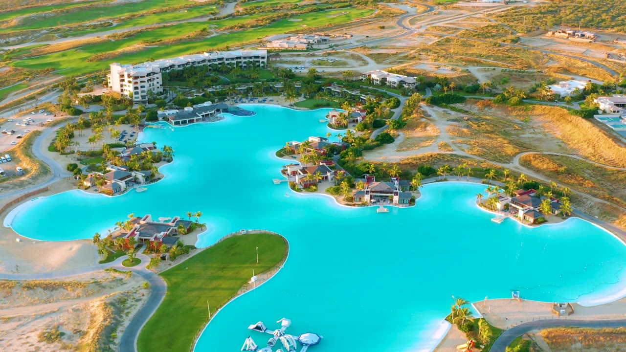 Aerial view over Diamante Cabo San Lucas showing the turquoise man-made lagoon, clusters of houses with palms, pale sandy banks, a green lawn area, and no visible people.
