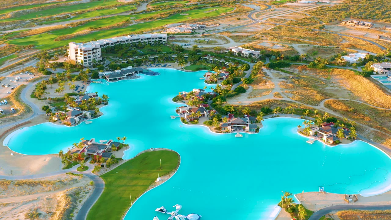Wide aerial composition of lagoon, homes, green grass, and sandy desert roads.