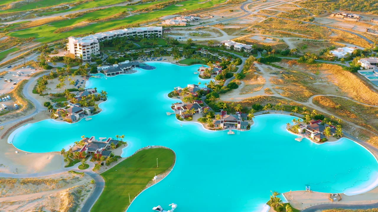 Aerial view over Diamante Cabo San Lucas showing a turquoise artificial lake, island homes, curved sandy banks, golf-course grass, dry land, and no visible people.