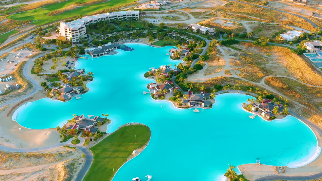 Opening aerial frame over a turquoise lake at Diamante Cabo San Lucas, with residential buildings near the top edge, curved roads, green lawns, sandy banks, and no visible people.