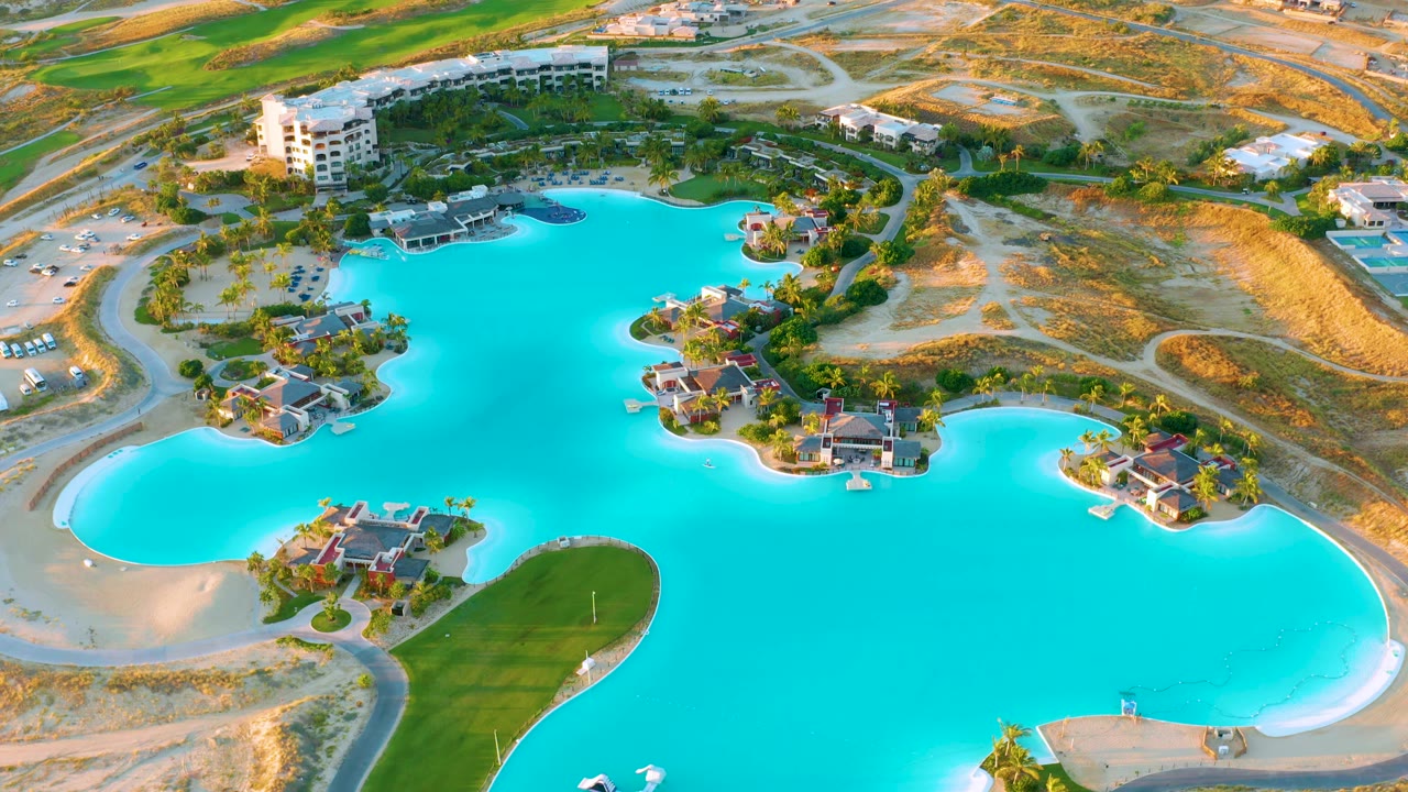 Overhead view of bright blue water with homes, roads, palms, and sandy terrain around it.