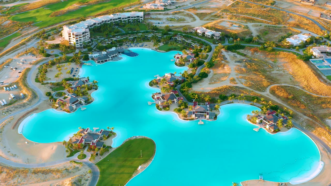 Aerial frame from Diamante Cabo San Lucas showing turquoise lagoon water surrounding small residential islands, pale sand edges, green grass, roads, resort buildings, and no visible people.