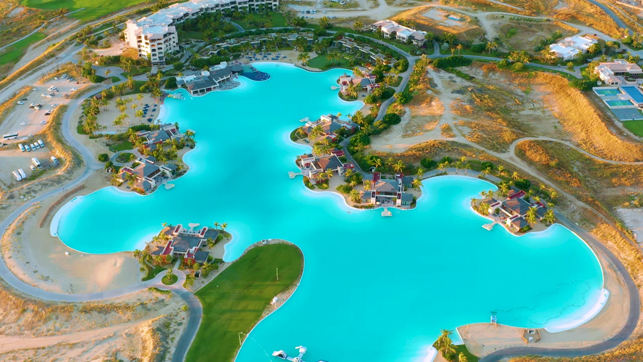 Aerial view at 0:01 showing bright blue lake water wrapping around small residential islands at Diamante Cabo San Lucas, with exposed sandy shore, roads, dry hillsides, and no visible people.