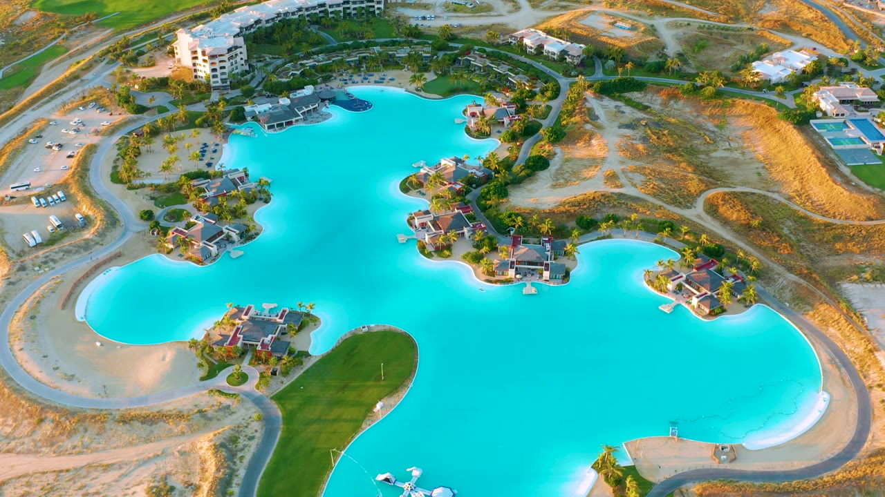 At 0:02, the high aerial frame shows the man-made lake at Diamante Cabo San Lucas with island homes, a curved green lawn area, road loops, dry lakebed edges, and no visible people.
