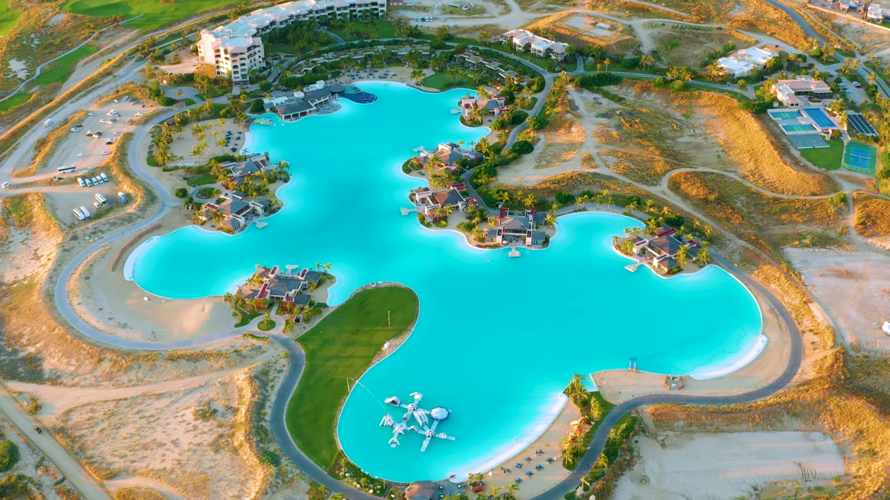 High drone frame of the Diamante Cabo San Lucas lagoon, with the green peninsula lower in frame, sandy lots to the sides, villas along the water, and no visible people.