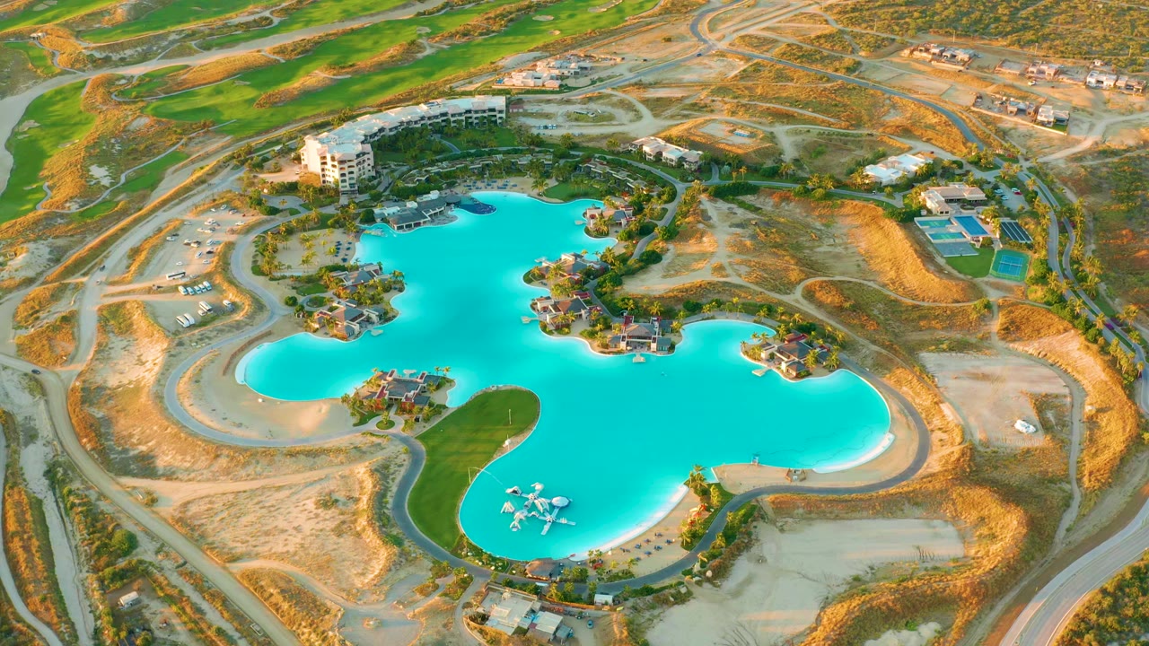 Second aerial frame of the Diamante Cabo San Lucas reservoir, showing the turquoise water centered between small landscaped islands, roads, buildings, dry land, and no visible people.