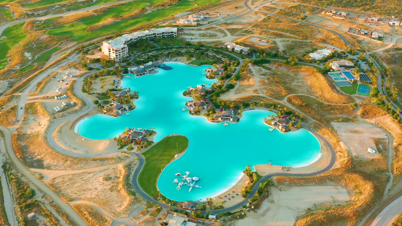 High aerial view over a turquoise artificial lake at Diamante Cabo San Lucas, with curved sandy banks, green grass, roads, buildings, dry lots, and no visible people.
